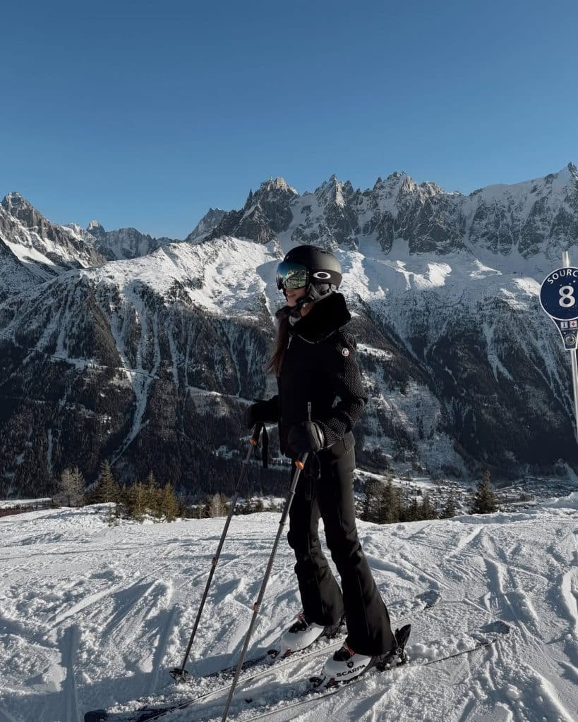 Woman in full black ski gear posing on a mountain slope under clear blue skies.