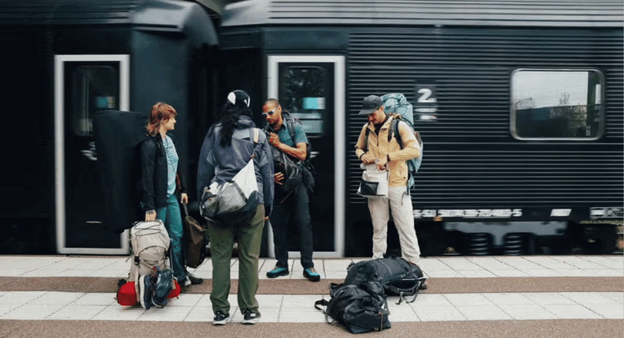 Travelers with backpacks and layered outdoor jackets waiting at a dark train station platform.