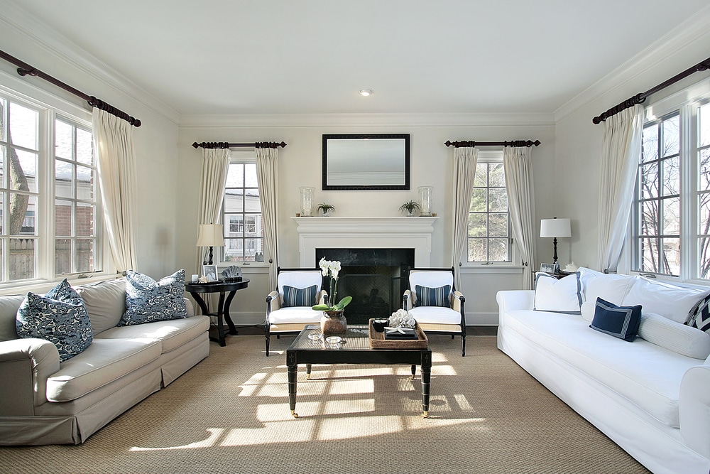 An elegantly furnished living room with white sofas, blue patterned pillows, and large windows providing natural light.