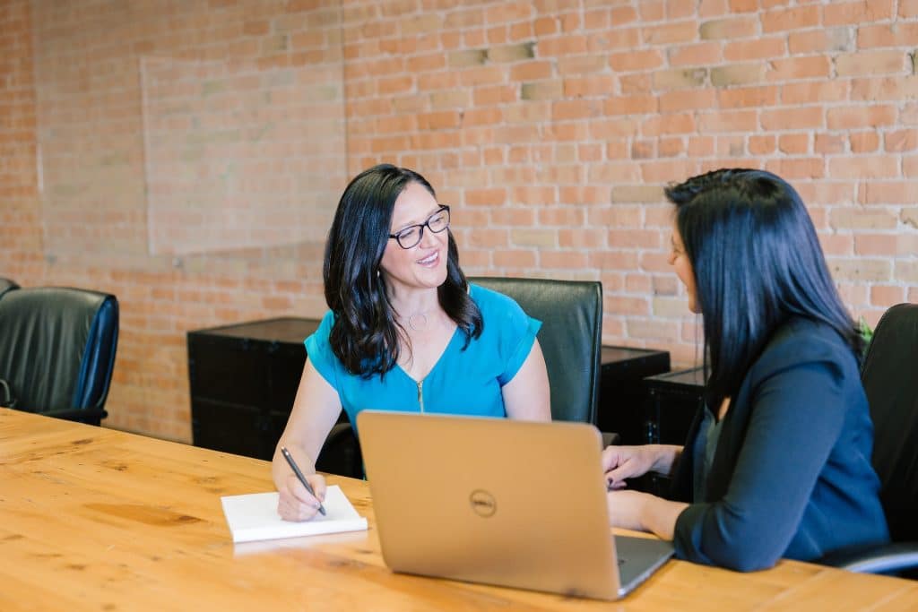 Two women in professional attire, one in a teal V-neck and the other in a blazer, using a laptop in a modern office.