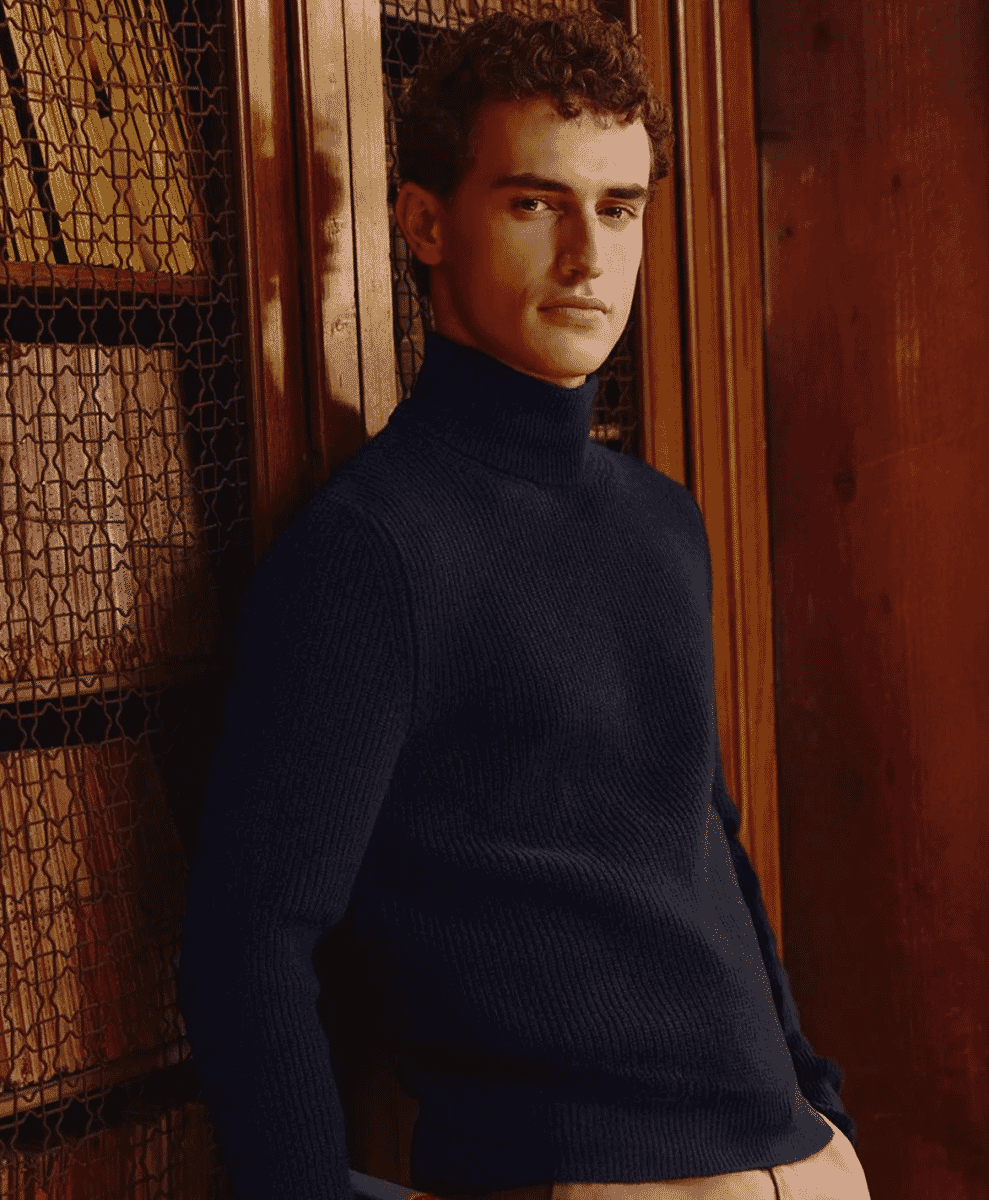 A man wearing a navy blue ribbed turtleneck standing against a wooden bookshelf.
