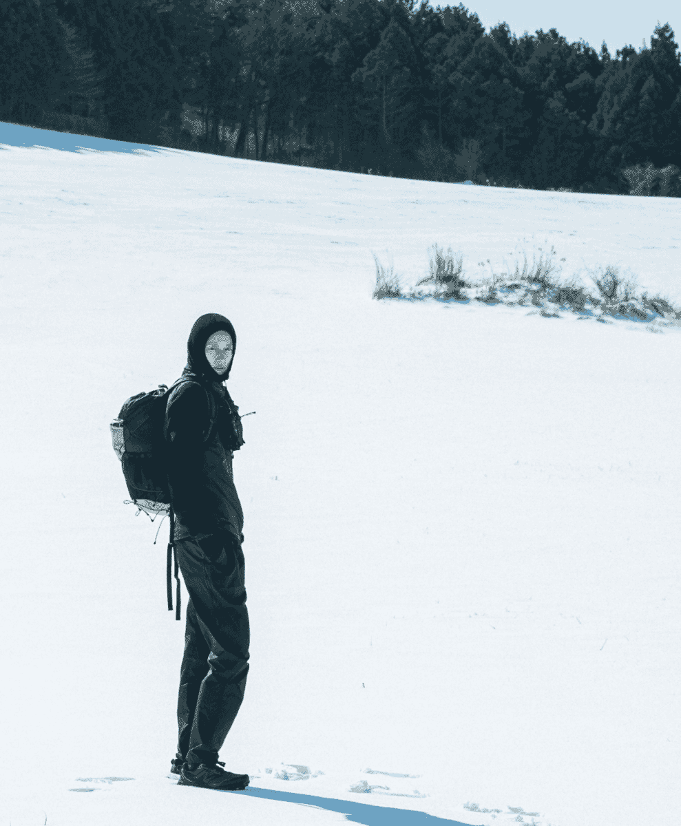 A person in full black technical outdoor gear and a backpack standing on a snowy hillside