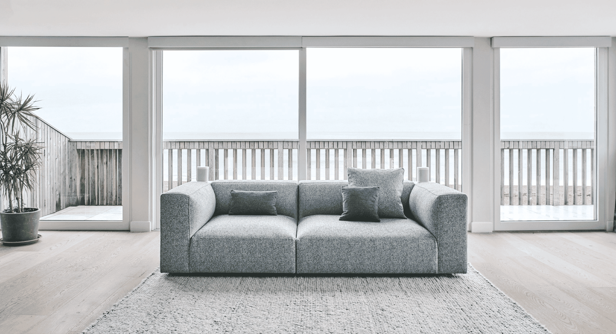 Modern minimalist living room with a gray textured sofa, wooden flooring, and a seaside balcony view.