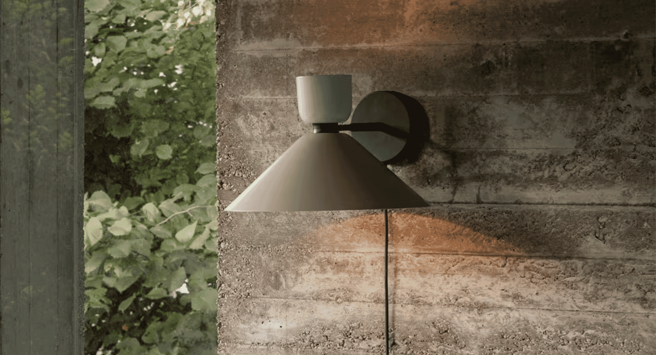 Industrial-chic wall sconce on a concrete wall beside green foliage seen through a window.