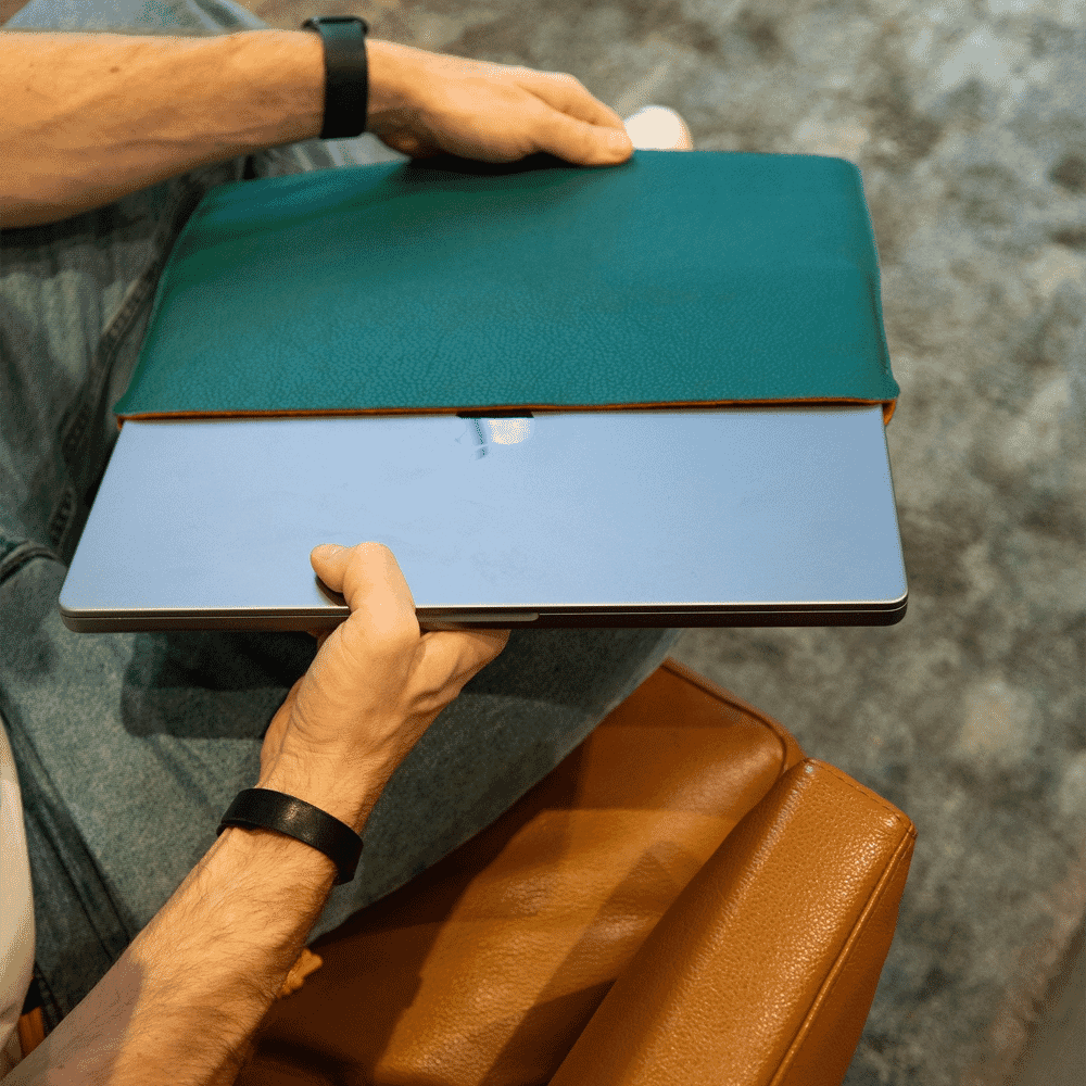 A person holding a teal leather laptop sleeve over a silver laptop in a relaxed seating area.