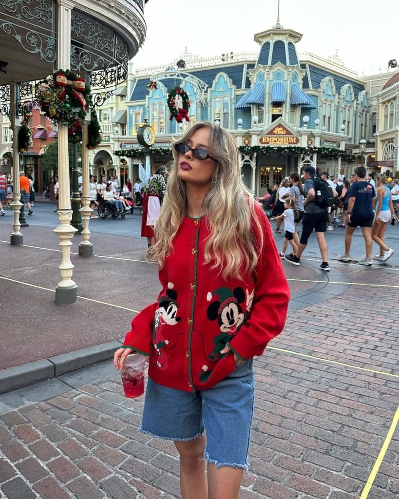 A woman in a red Mickey Mouse Christmas sweater and denim shorts on a festive street.