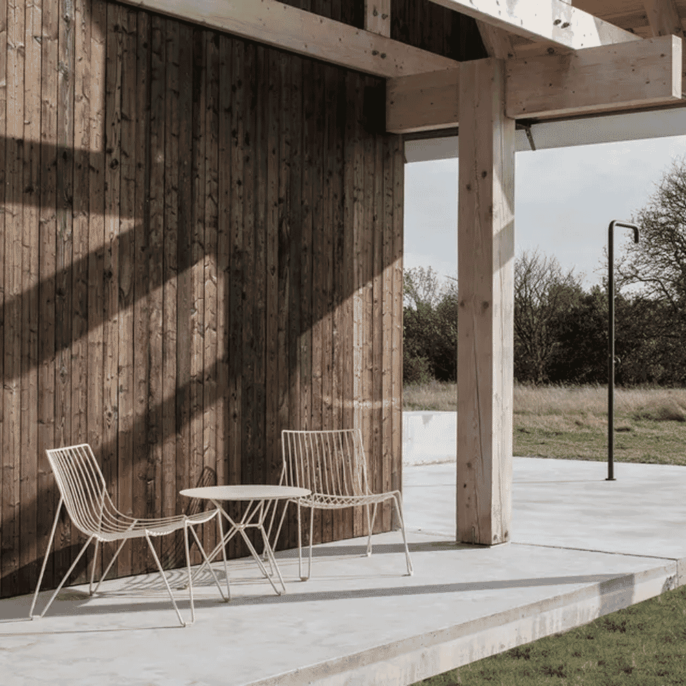 White wireframe chairs and a small table on a concrete patio under a modern timber-framed roof.