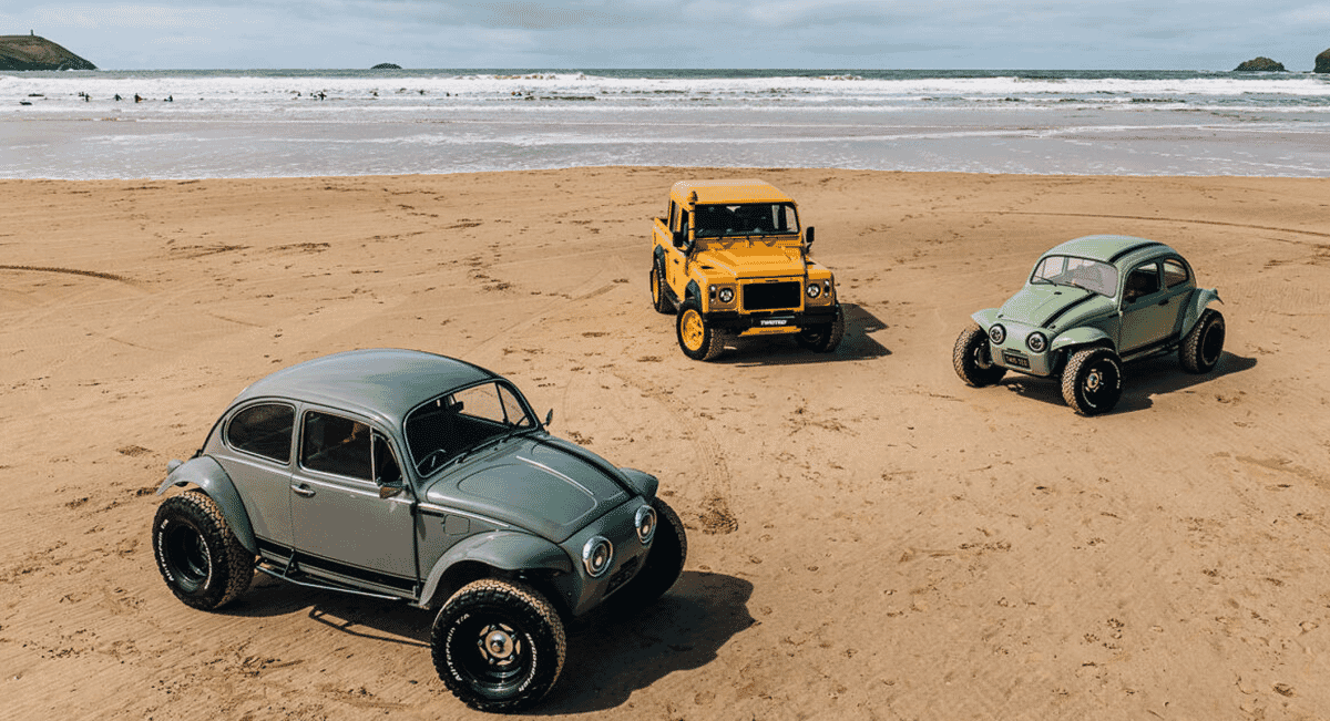 Two customized Beetles and a Land Rover parked on a sandy beach near ocean waves.