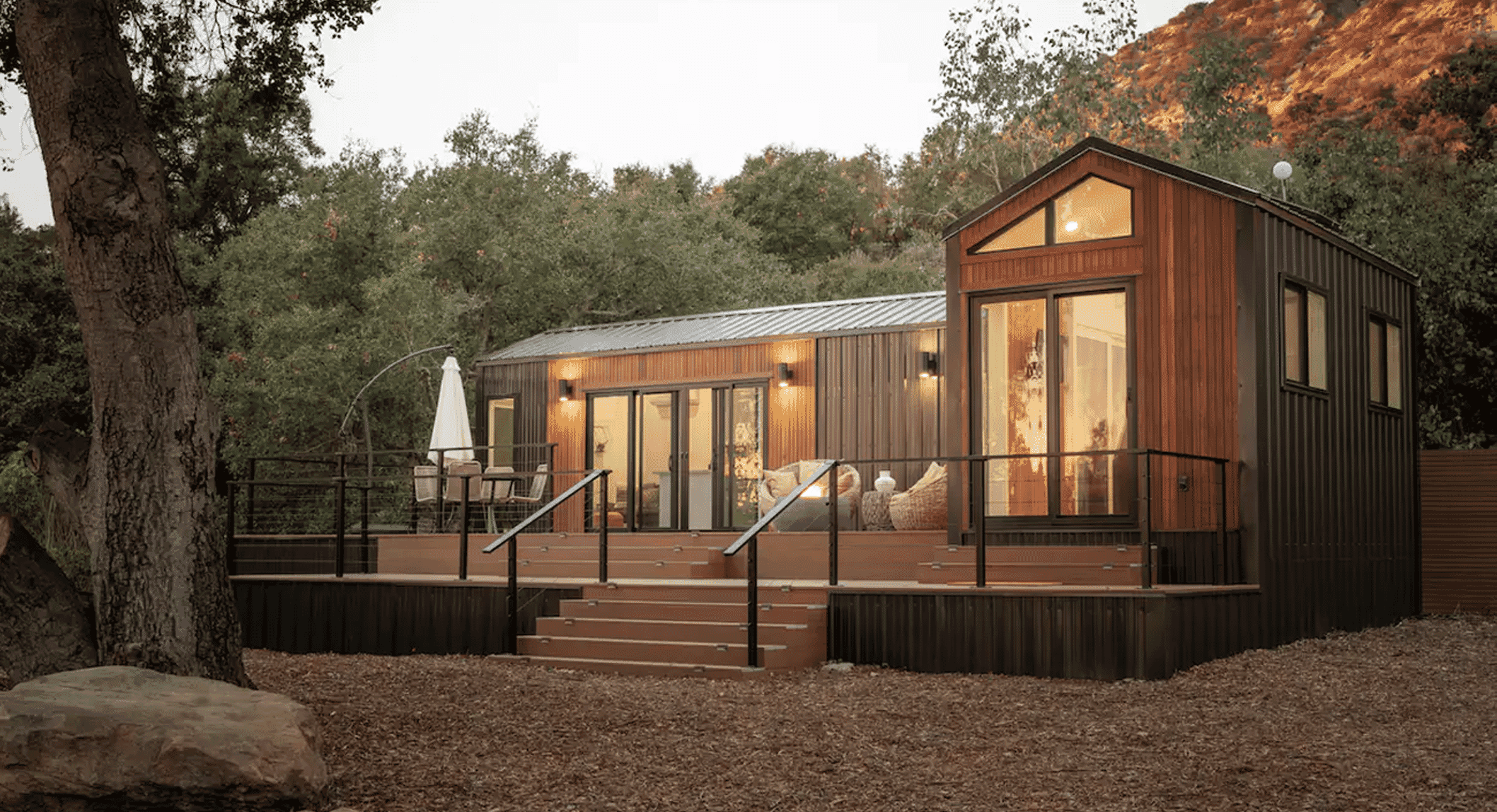 A modern tiny house with wood siding and large glass windows illuminated at dusk.