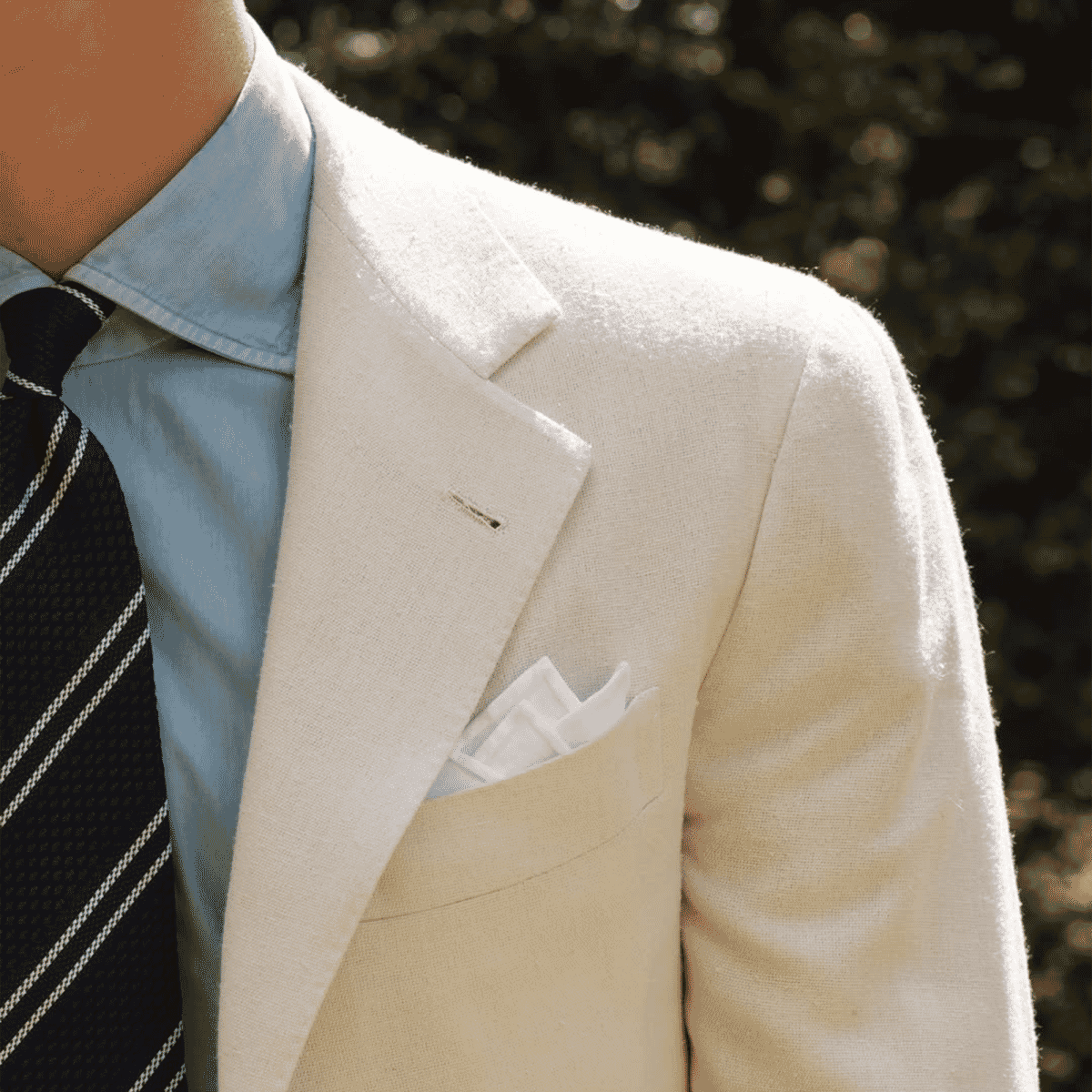 Close-up of a man wearing a light beige textured blazer, light blue shirt, and a striped tie with a white pocket square.