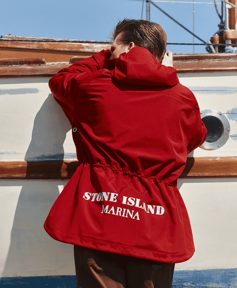 A man in a red hooded Stone Island Marina jacket standing on a boat deck at sea.