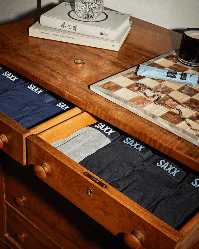 Neatly folded navy, gray, and black SAXX boxer shorts in a wooden dresser drawer alongside lifestyle accessories.