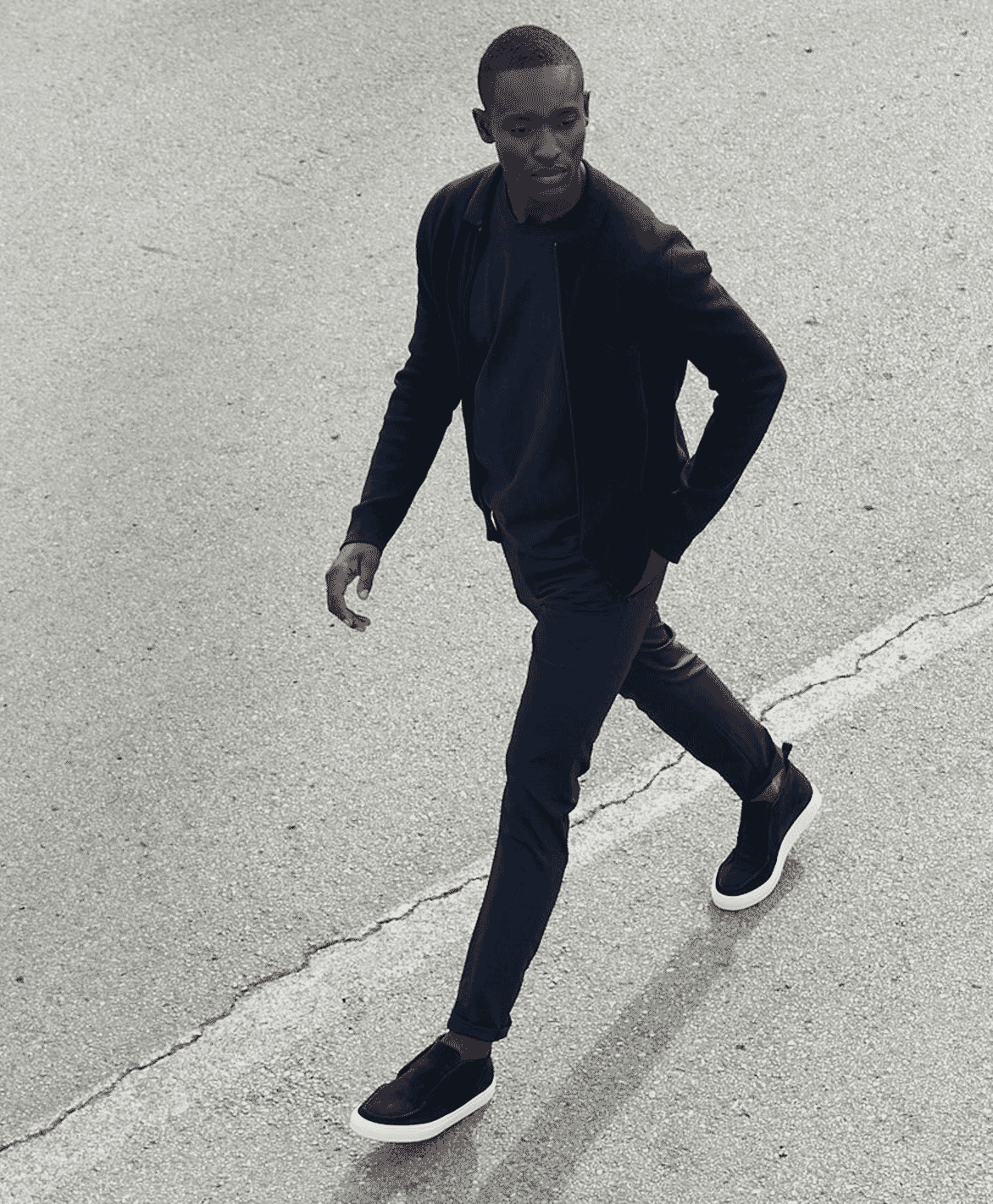 Man in all-black attire and slip-on sneakers walking across an asphalt street.