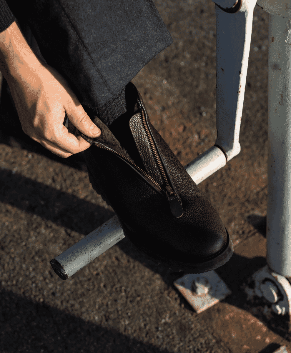 Person in dark wool trousers adjusting a black leather zip boot against a city pavement backdrop.