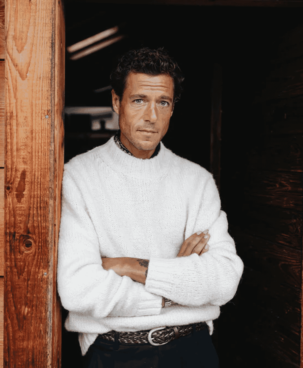 Man in a white chunky knit sweater standing in a rustic wooden doorway.