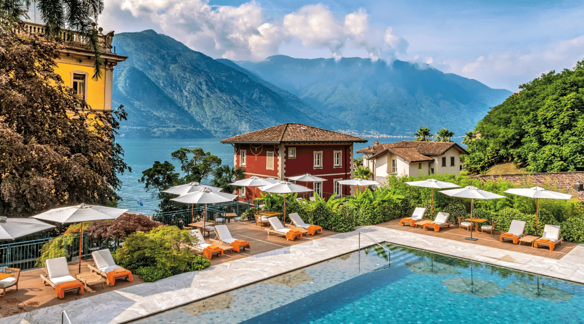 A luxury lakeside resort with orange lounge chairs and white umbrellas against a mountain backdrop.