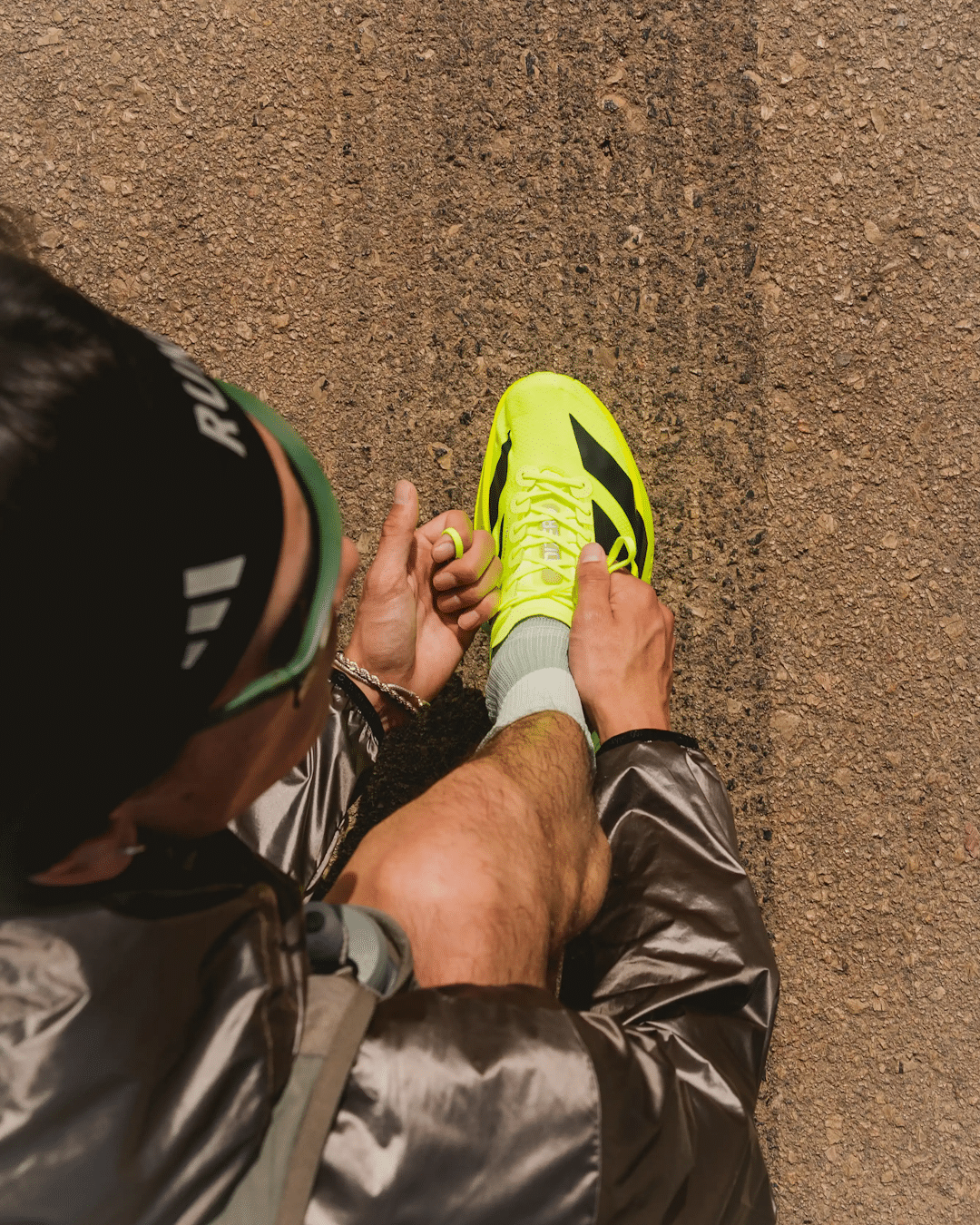 A runner in a silver jacket and neon yellow shoes adjusting laces on a gravel path.