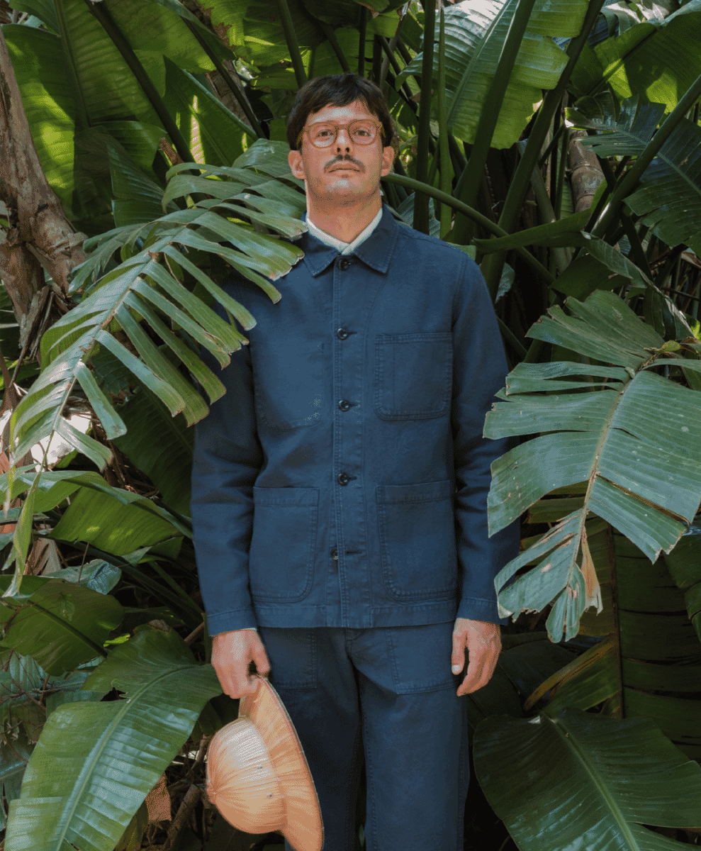 Man in a navy work jacket and trousers holding a straw hat in a tropical garden