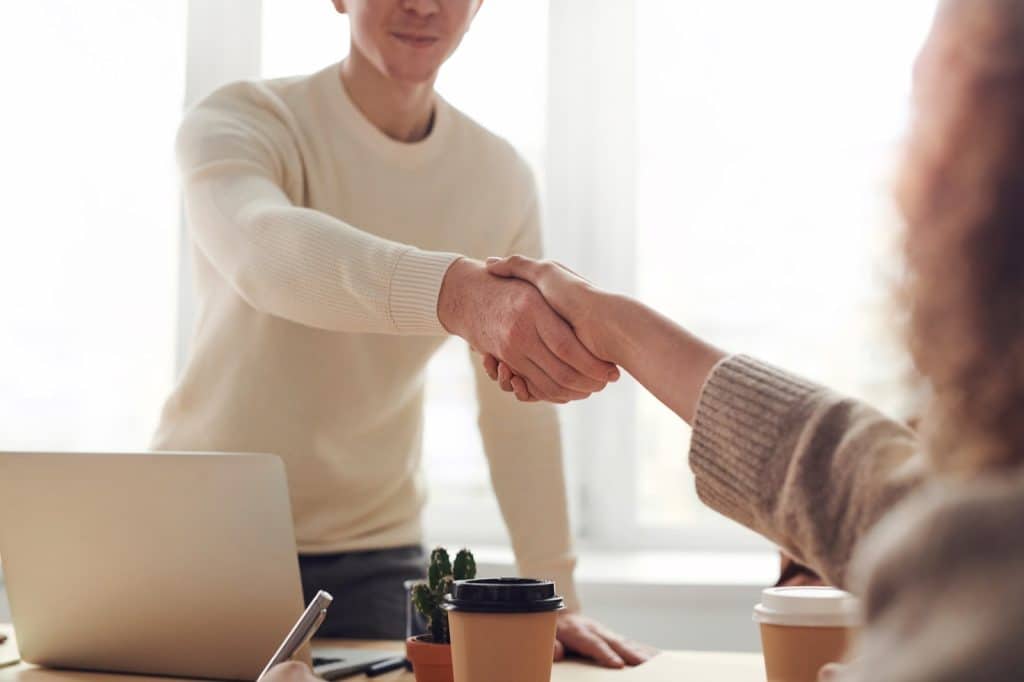 Two professionals in knit sweaters shaking hands across a table in a bright office.