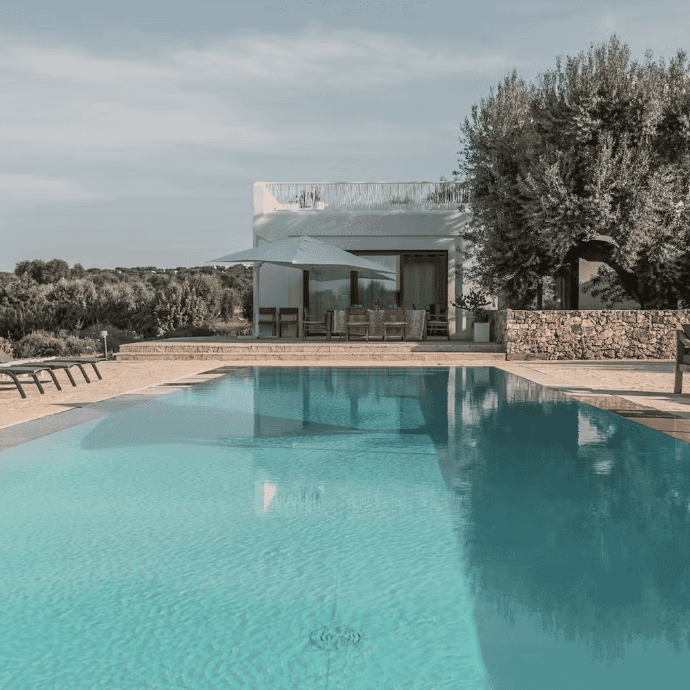Serene modern villa with a turquoise swimming pool and olive trees under a soft sky.
