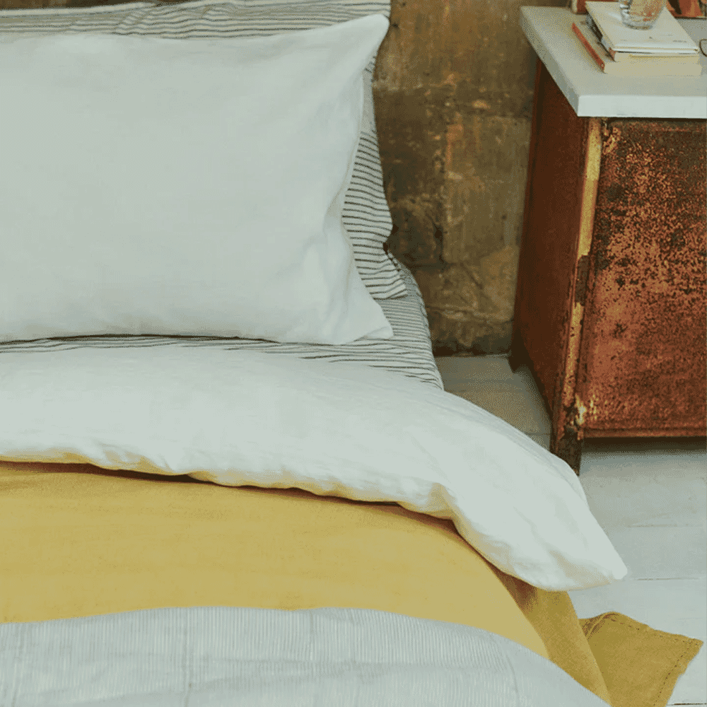 A cozy bed with white and yellow linen sheets and a rustic nightstand.