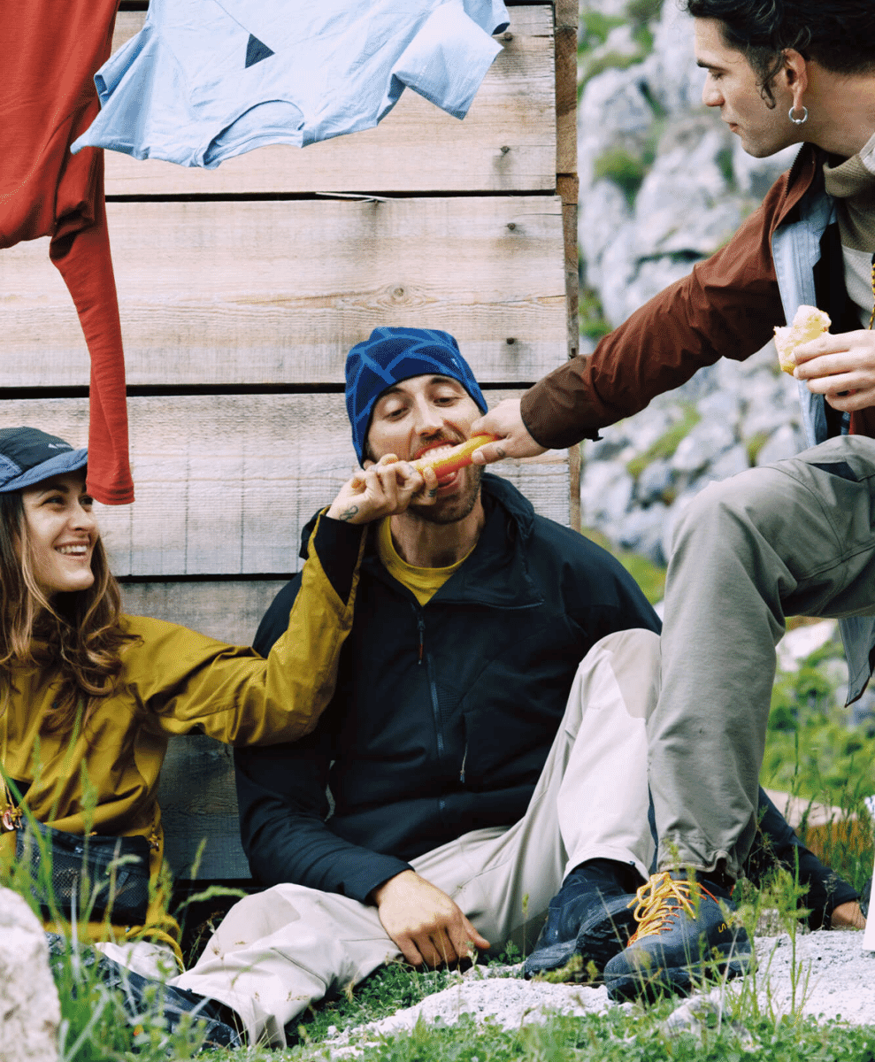 A group of three hikers in layered outdoor wear sharing food outside a rustic wooden cabin in the mountains.