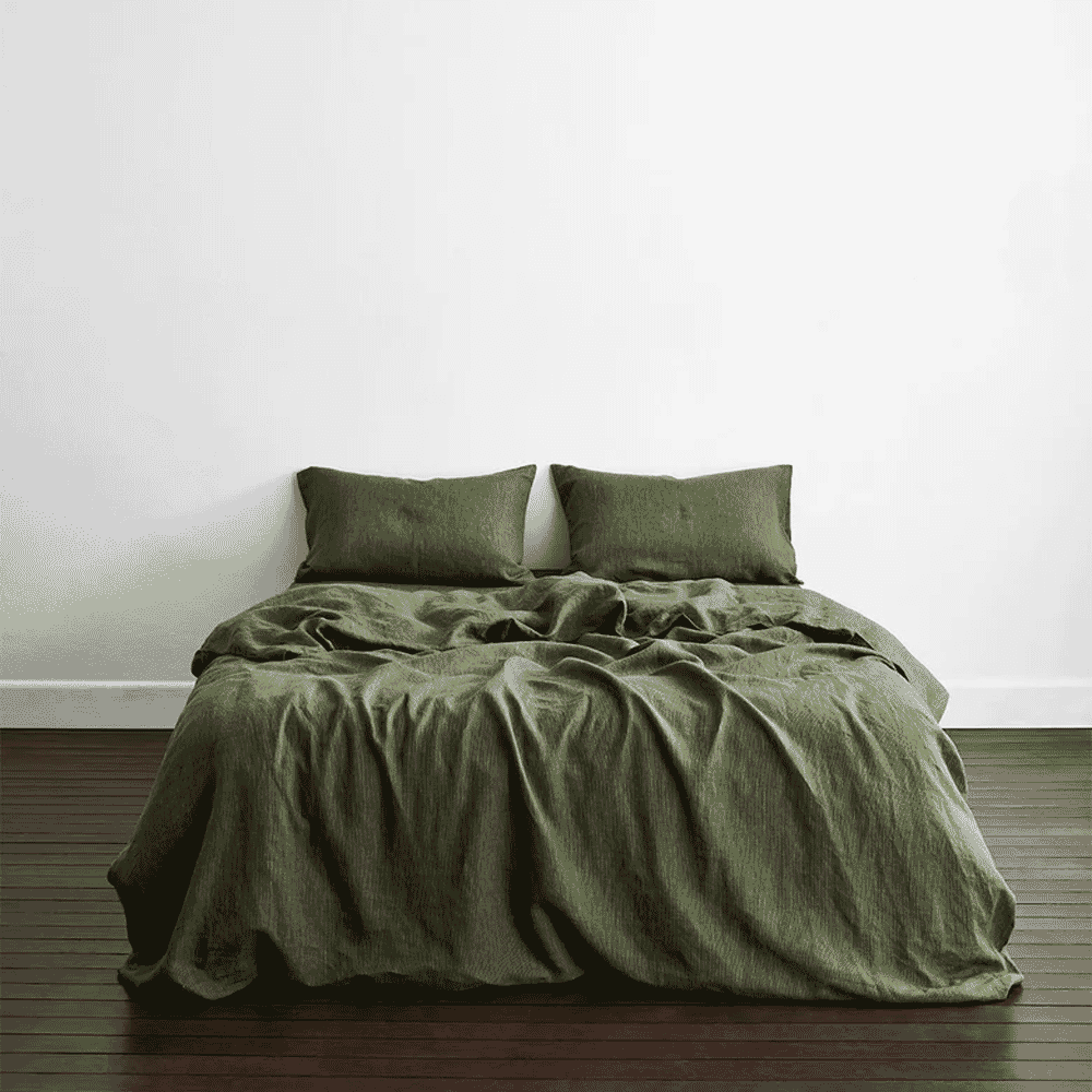 A bed dressed in olive green linen bedding against a minimalist white wall.
