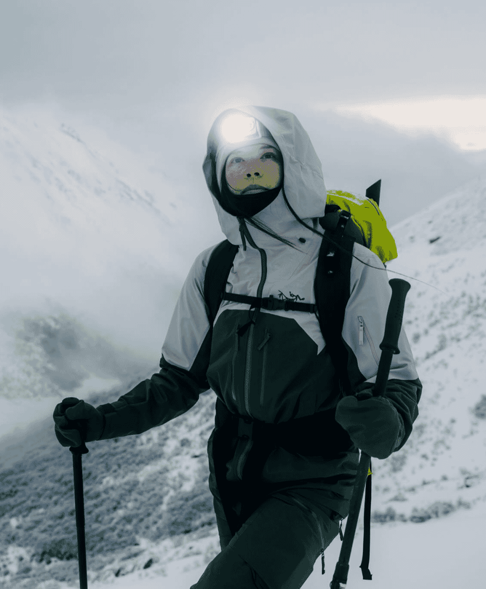 A hiker with trekking poles and a white insulated jacket navigating a snowy mountain landscape.