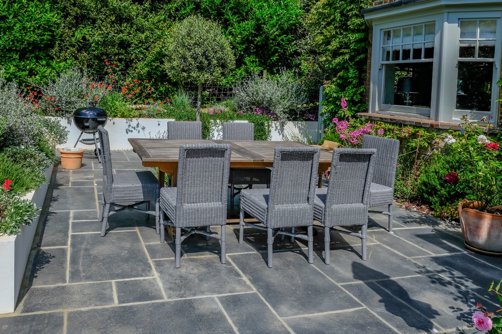 Sunlit patio with a wooden dining table and grey wicker chairs set against slate stone paving and flowering garden beds.