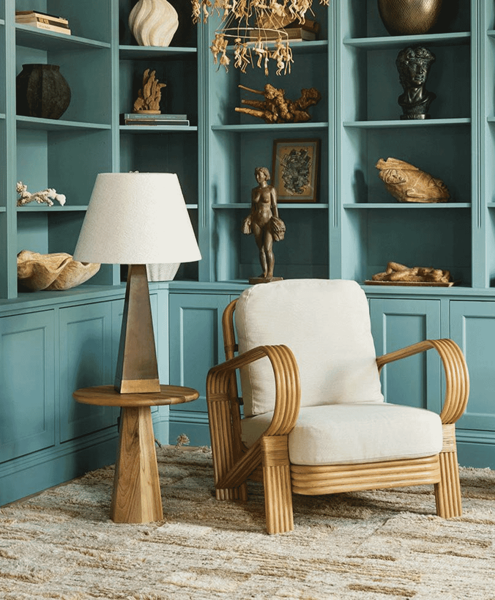 A mid-century modern rattan lounge chair with cream upholstery in a blue-walled room with decorative sculptures.