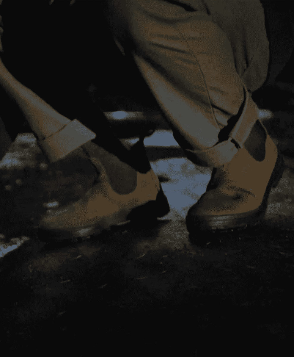 Close-up of a person in beige trousers and brown Chelsea boots on a gritty urban pavement.
