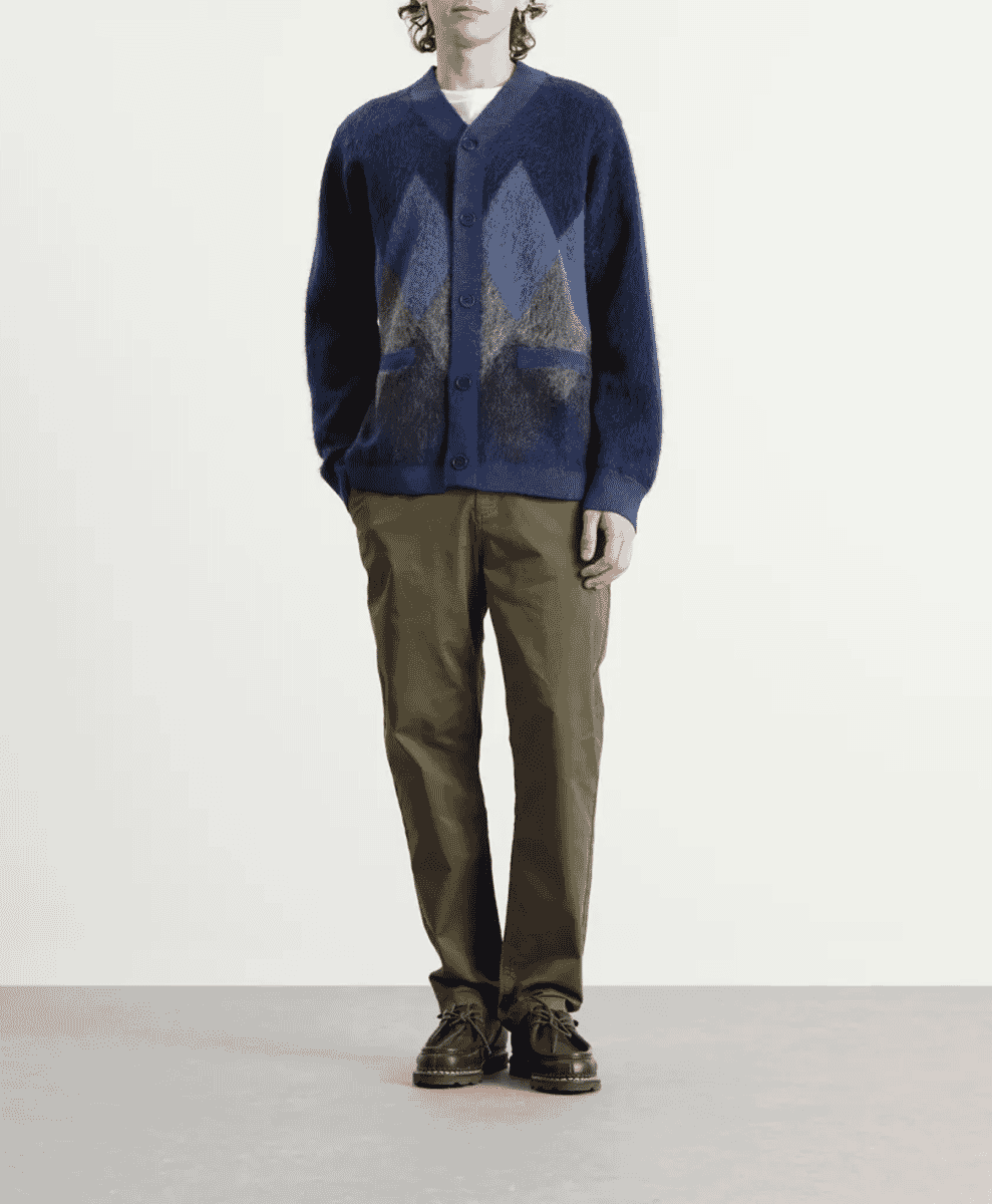 A model in a navy blue argyle cardigan with geometric patterns, olive trousers, and leather shoes.
