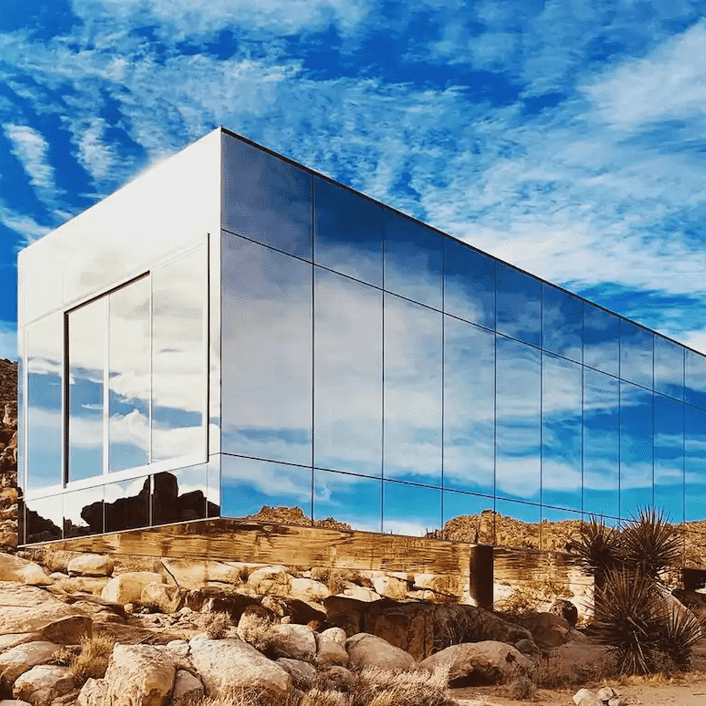 A modern mirrored cube structure reflecting the desert sky and rocky terrain.