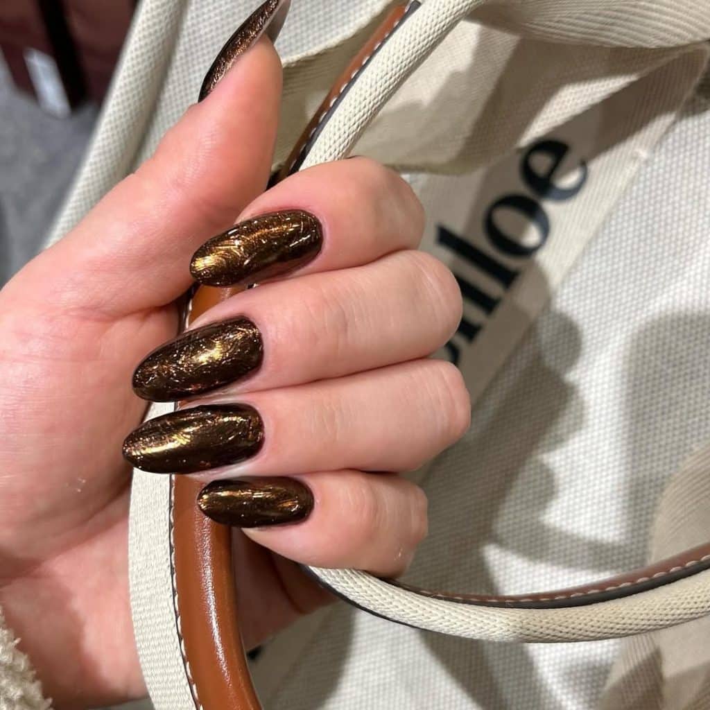 Metallic bronze long nails holding a beige Chloé leather handbag.