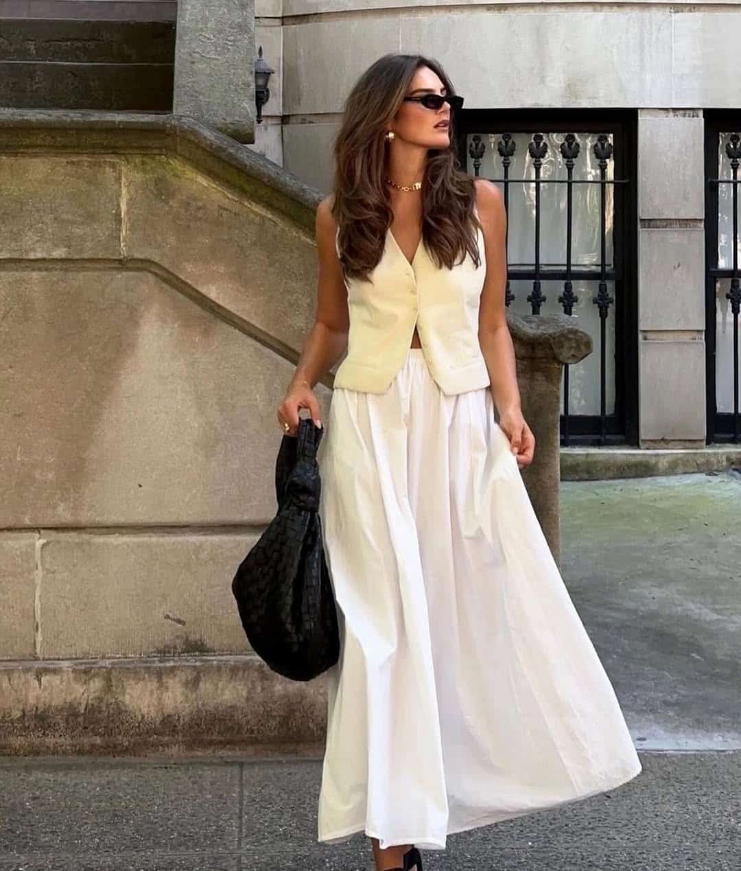 A woman in a cream sleeveless vest and white maxi skirt with black sunglasses on a city sidewalk.