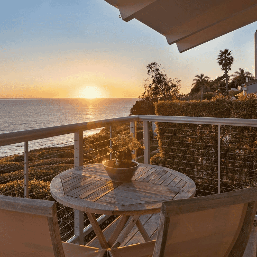 A serene ocean sunset viewed from a wooden balcony with a round table and hedges.