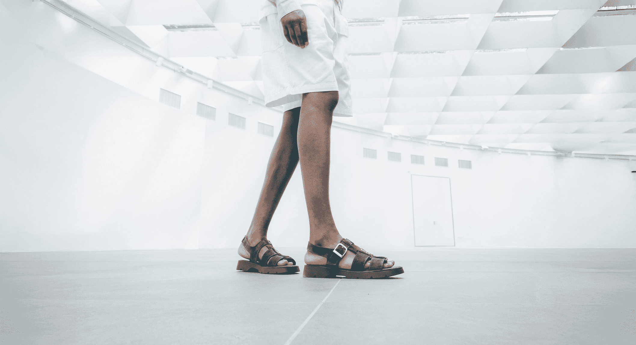 Full-body shot of a person in a white minimalist outfit wearing chunky brown leather sandals.