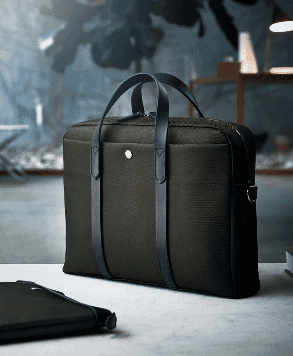 An olive green and black leather-trimmed briefcase sits on a marble desk in a modern office.