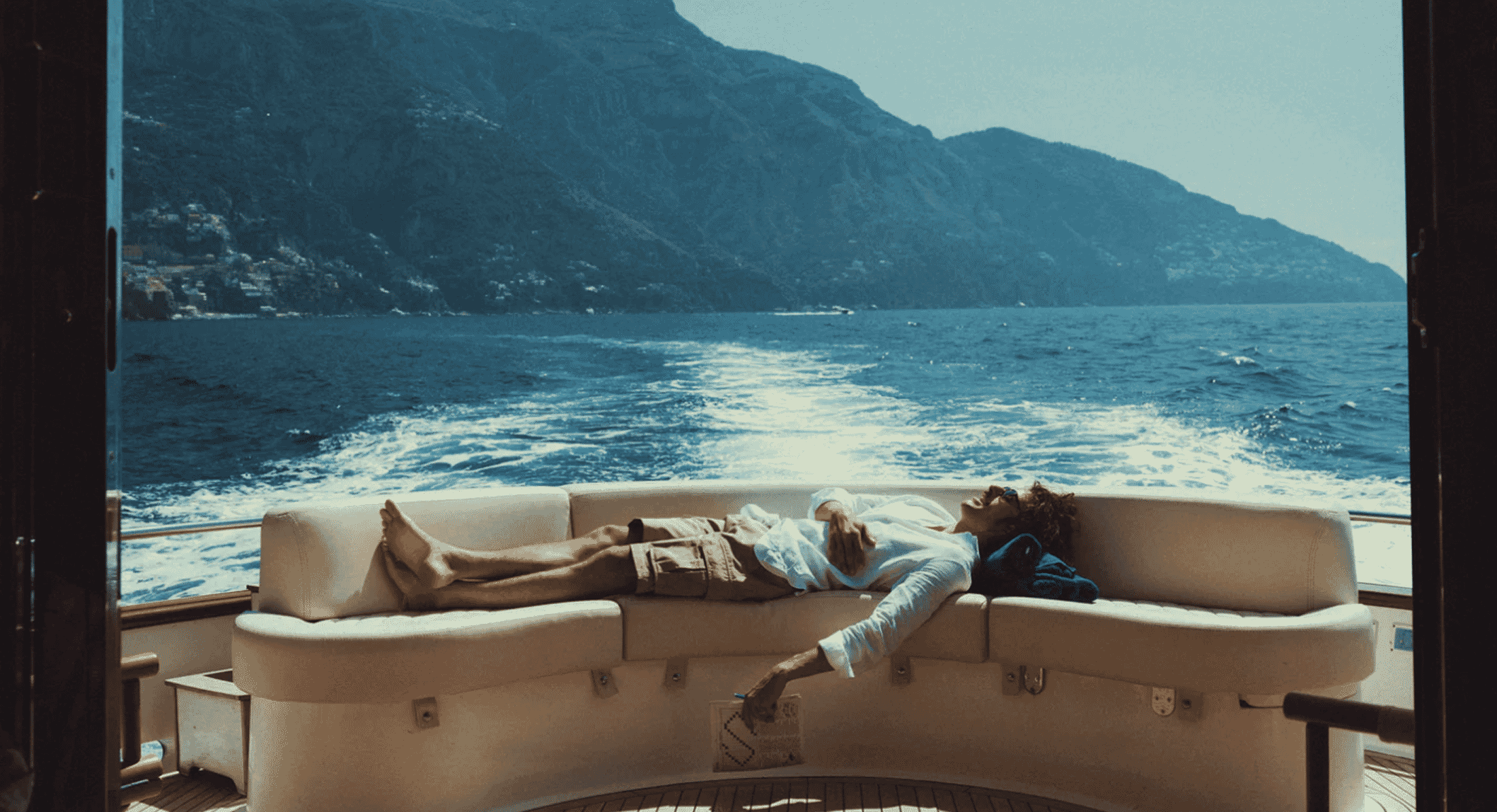 A man wearing a light blue linen shirt and beige shorts lounging on a white yacht sofa.