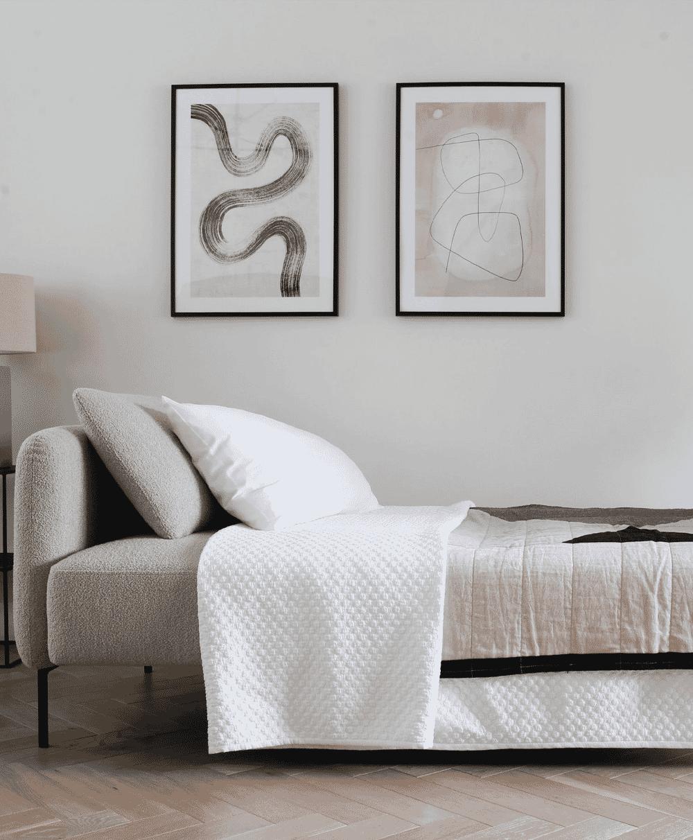 Minimalist bedroom setting with a gray sofa bed, white bedding, and abstract wall art.