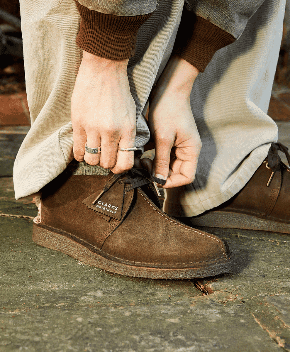 Close-up of a person tying the laces of brown suede desert boots on a stone path.