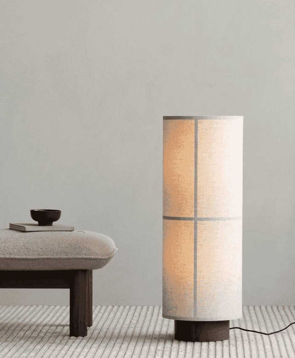 A cylindrical floor lamp with a textured fabric shade standing on a striped rug against a neutral wall.
