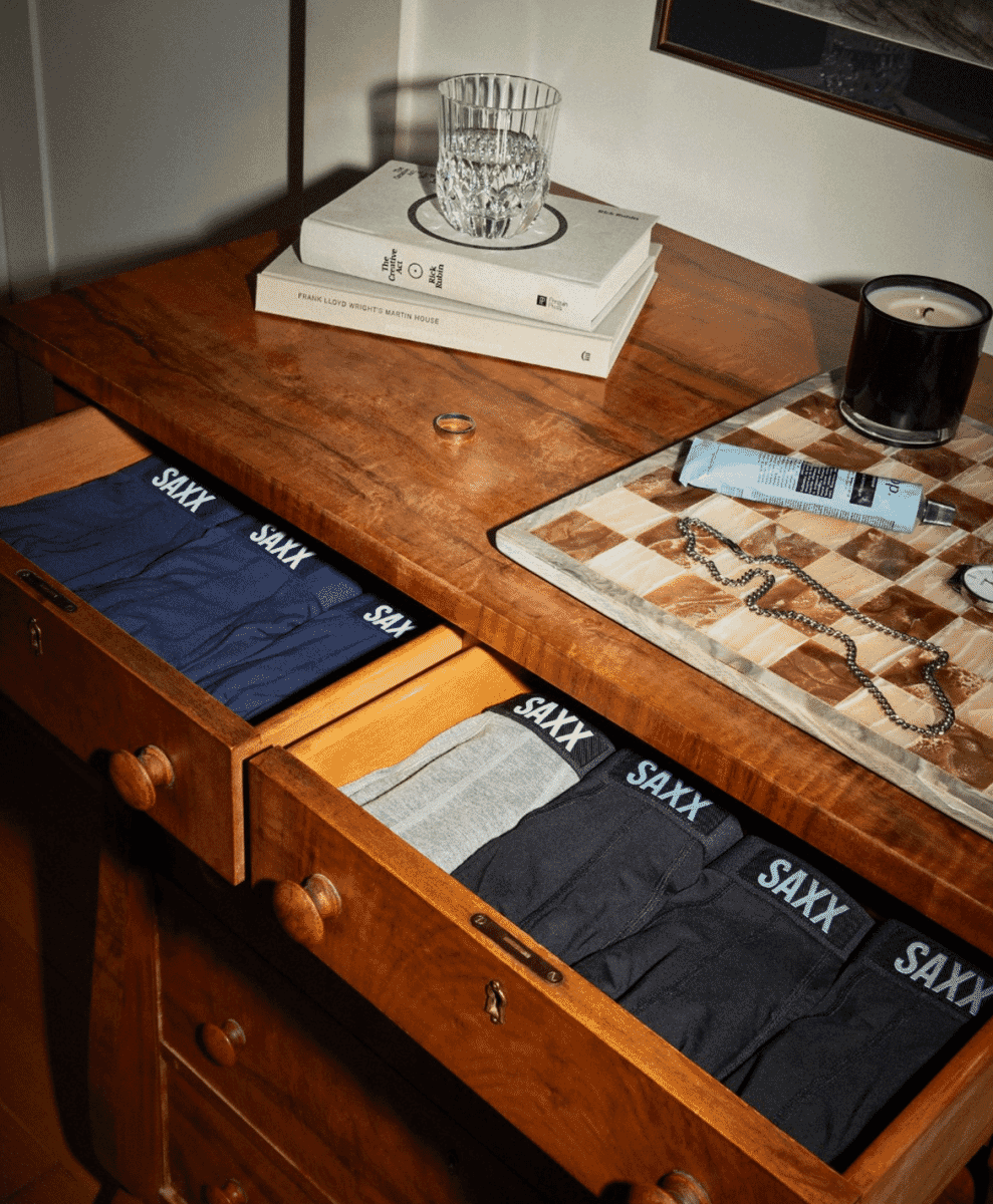 SAXX underwear neatly folded in a dark wooden dresser drawer next to lifestyle items like a candle and glass.