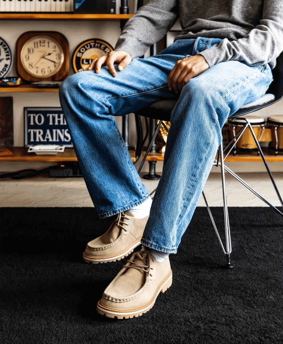 Man in a cozy vintage room wearing beige suede moccasins with white soles and denim jeans.