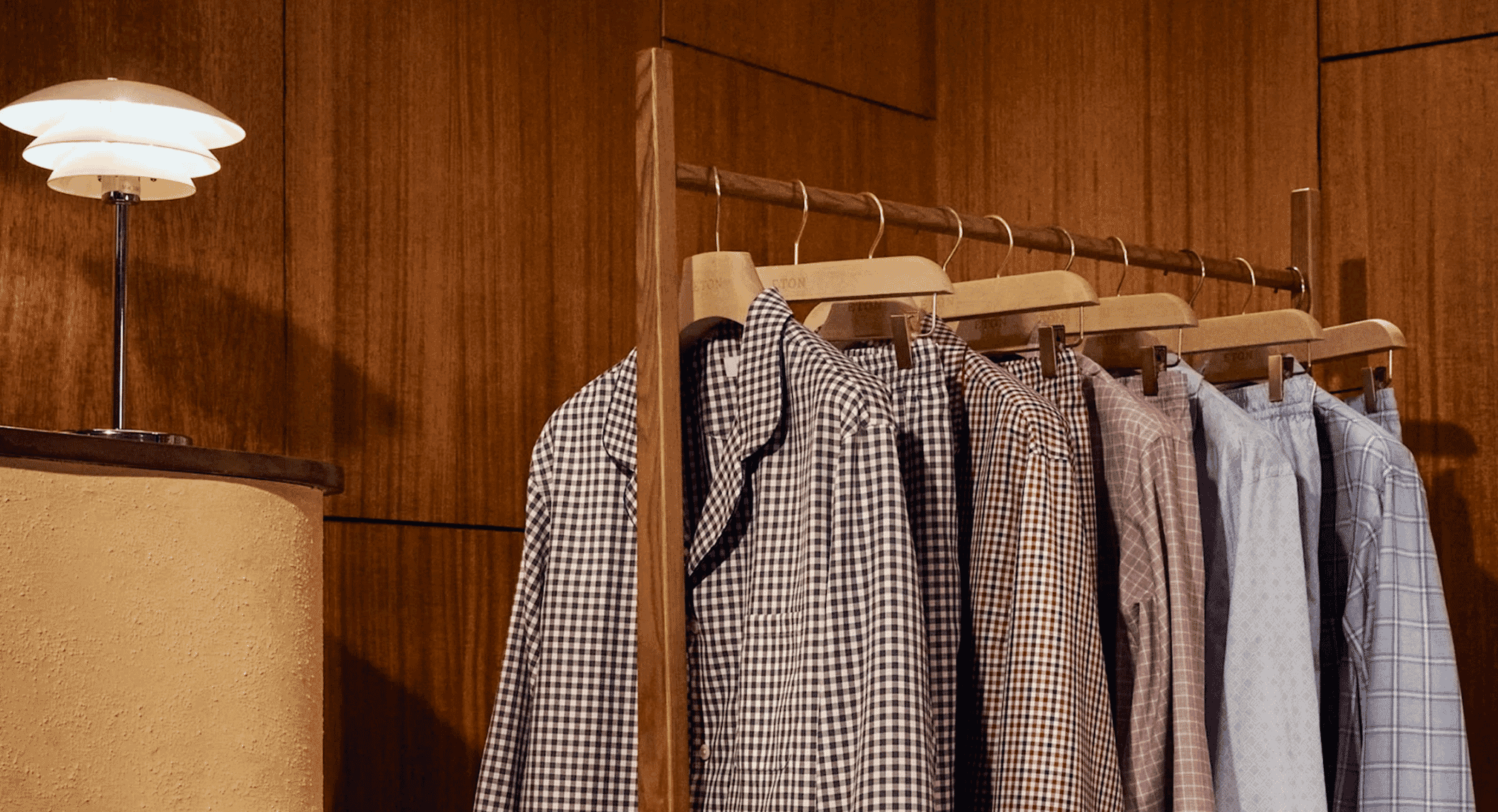 Men's checkered pajama sets hanging on a wooden rack in a warm, modern bedroom.
