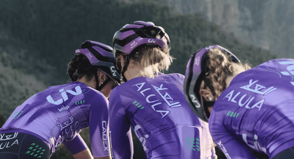 Three female professional cyclists huddling on a scenic mountain road in matching purple kits.