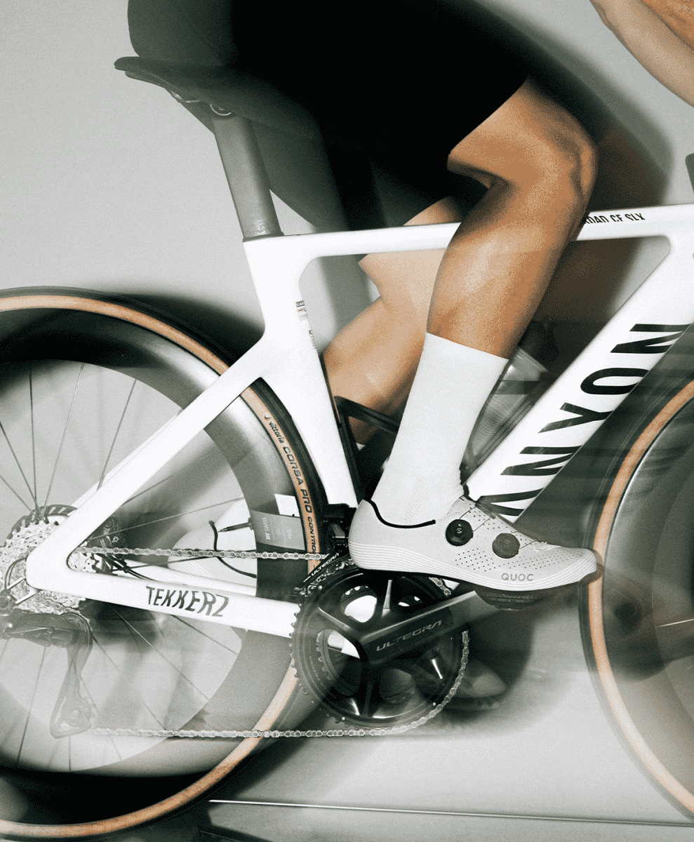 A cyclist on a white time-trial bike wearing white QUOC shoes and socks with a motion-blurred background.