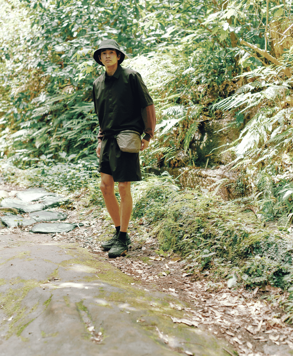 Man wearing a dark bucket hat, black technical shirt, and utility waist bag on a mossy forest trail.