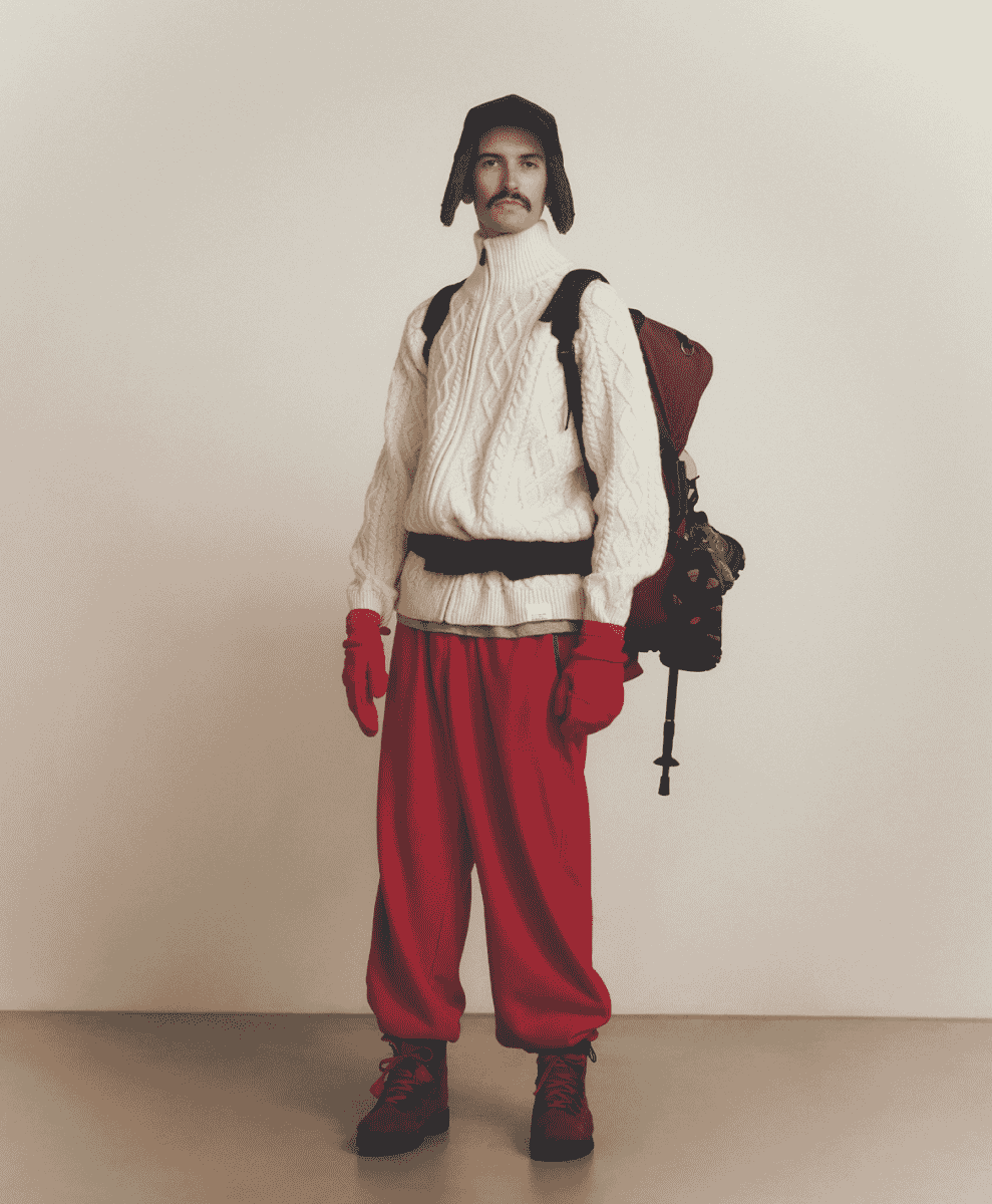 Man in a white cable-knit sweater, red baggy pants, and a black aviator hat.