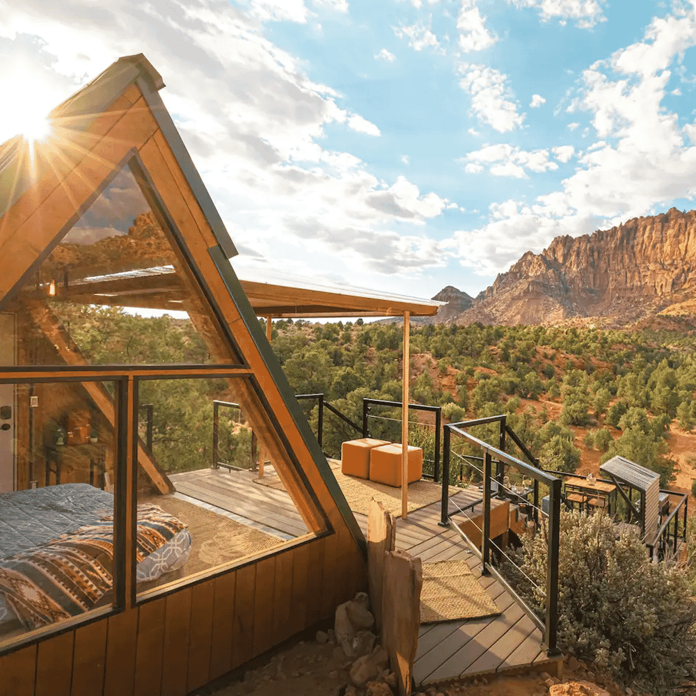 Modern A-frame cabin with glass walls overlooking a mountain valley at sunset.